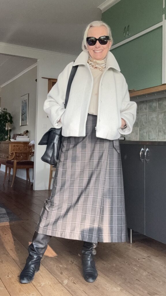 En kvinna med vitt hår bär solglasögon, en vit kavaj med krage, en beige blus med halsduk, en grårutig kjol och svarta stövlar. Hon står inomhus på ett trägolv, ler och har en svart väska över axeln.