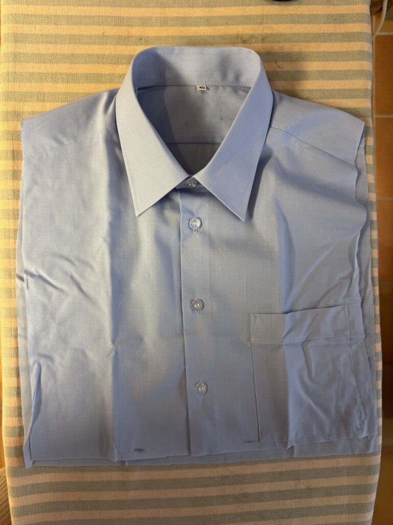 En prydligt vikt ljusblå skjorta med knappar och en framficka, placerad på en randig beige och grå yta.