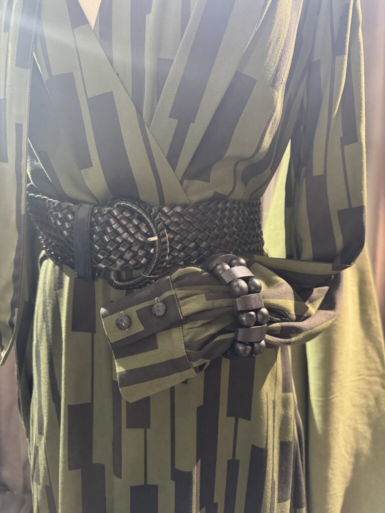 Närbild på en person som bär en grön och svart geometriskt mönstrad klänning med ett brett vävt svart bälte och ett tjockt svart armband över den manschettförsedda ärmen.