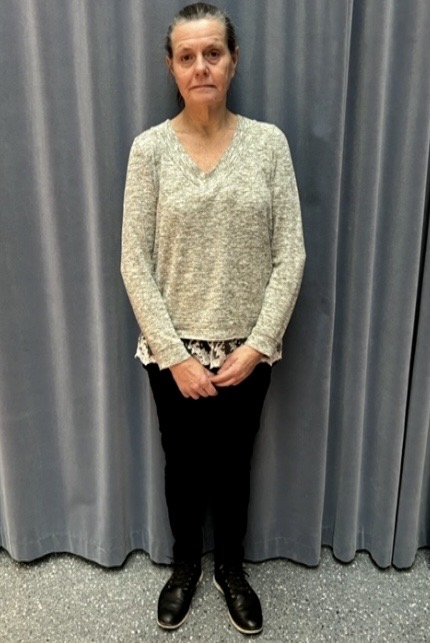 En person står inomhus framför gråa gardiner, klädd i en ljusgrå tröja över en mönstrad topp, svarta byxor och svarta skor, med händerna knäppta framför sig.
