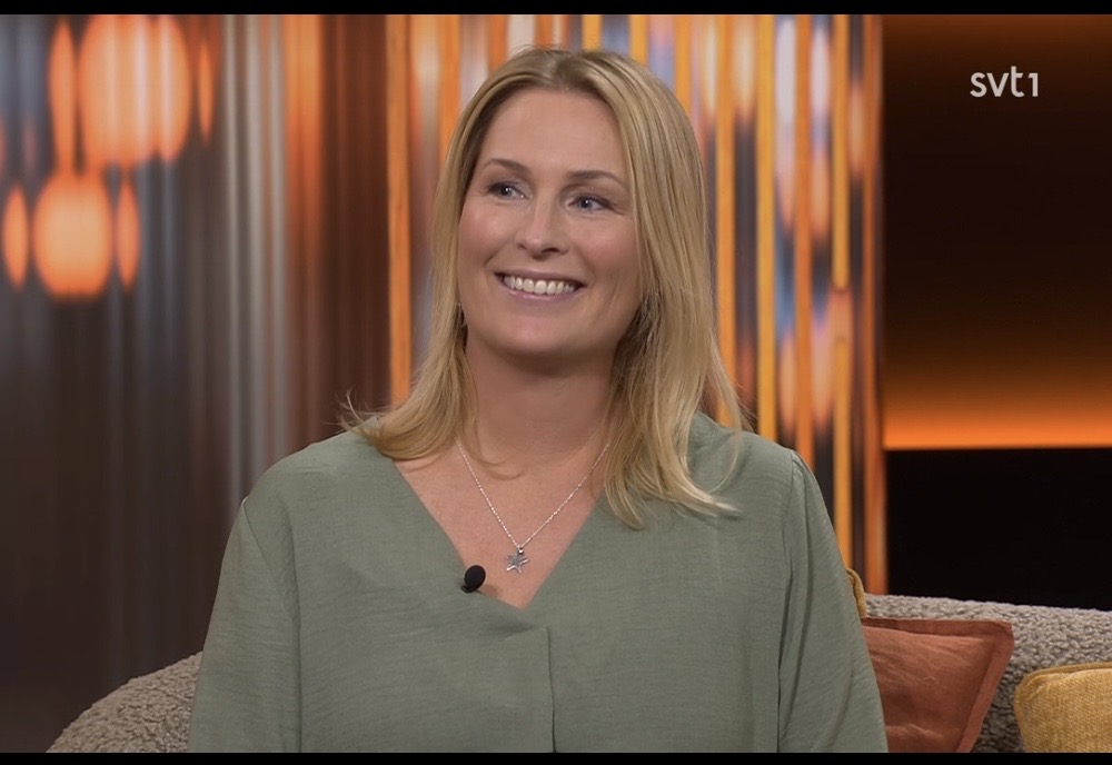 En kvinna med blont hår, iförd grön blus och halsband, ler när hon sitter i en soffa i en varmt upplyst TV-studio med orangea och bruna toner. SVT1:s logotyp syns i det övre högra hörnet.