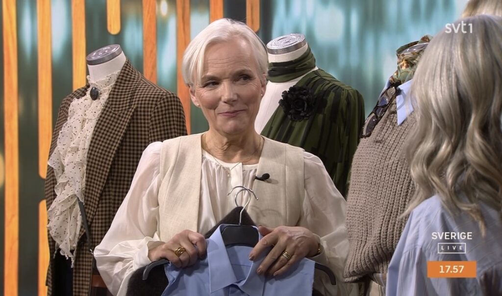 En kvinna med kort vitt hår håller en blå skjorta på en galge, stående bland skyltdockor klädda i vintagekläder, under en direktsändning på SVT med Sverige Live-logotypen och klockslag på skärmen.