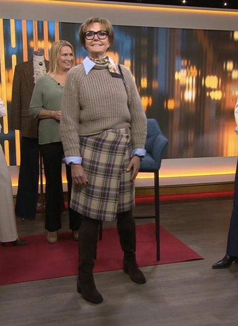 En kvinna med glasögon, en brun tröja över en blå skjorta, en rutig kjol, bruna stövlar och en halsduk poserar inomhus i en studiokuliss med suddiga ljus och människor i bakgrunden.