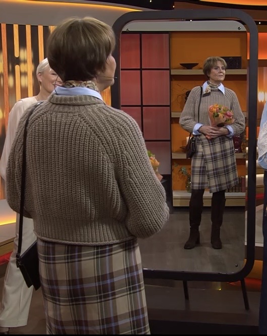 En kvinna i rutig kjol, ljusblå skjorta och brun stickad tröja står framför en hög spegel, håller i en bukett och tittar på sin spegelbild i ett varmt upplyst rum.