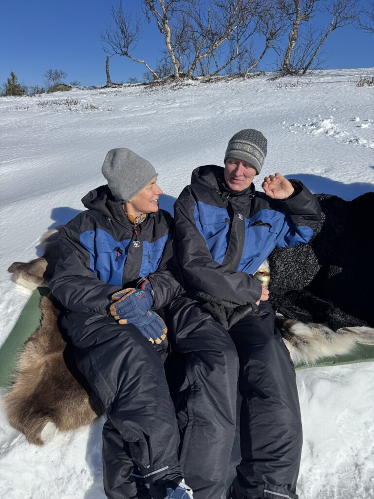 Två personer i blå och svarta vinterkläder sitter på pälsar i snön, ler och njuter av det soliga, snöiga landskapet. Lövlösa träd står bakom dem under en klarblå himmel.