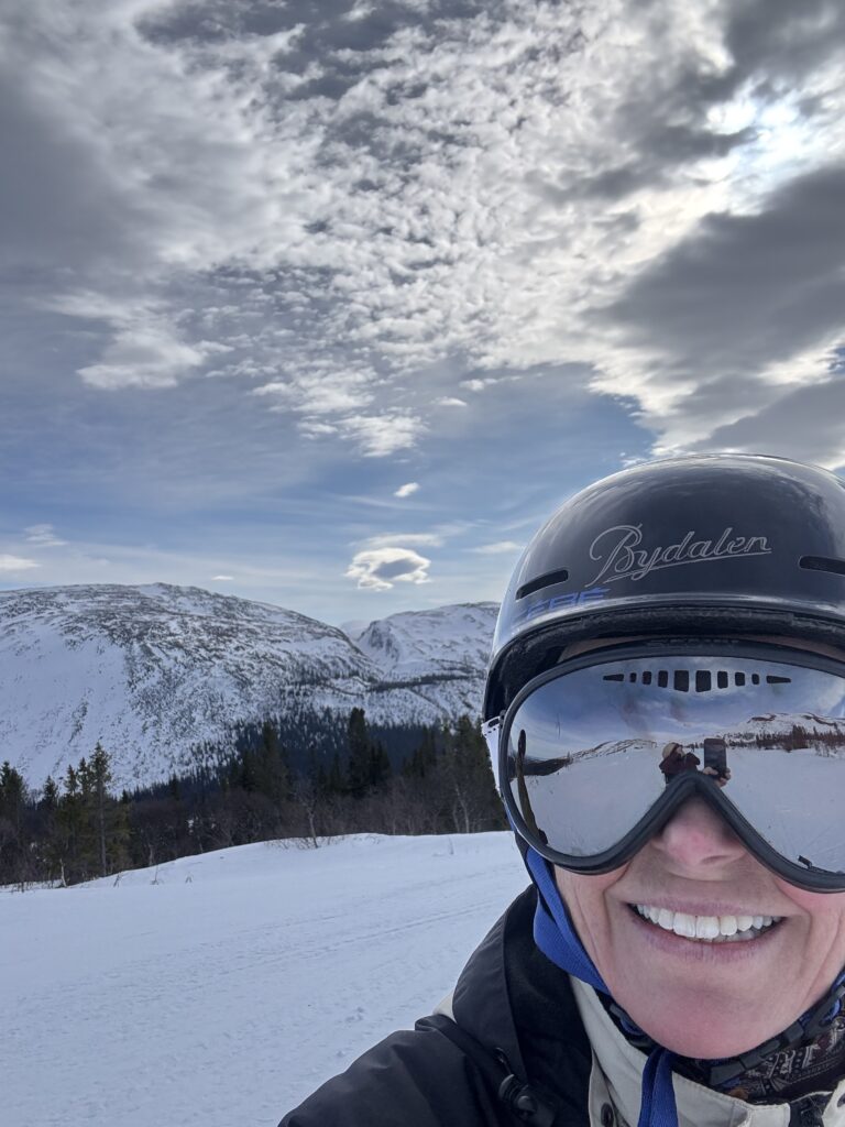 En person med hjälm och skidglasögon ler i förgrunden, med snöklädda berg, träd och en delvis molnig himmel i bakgrunden. På hjälmen står det "Bydalen".