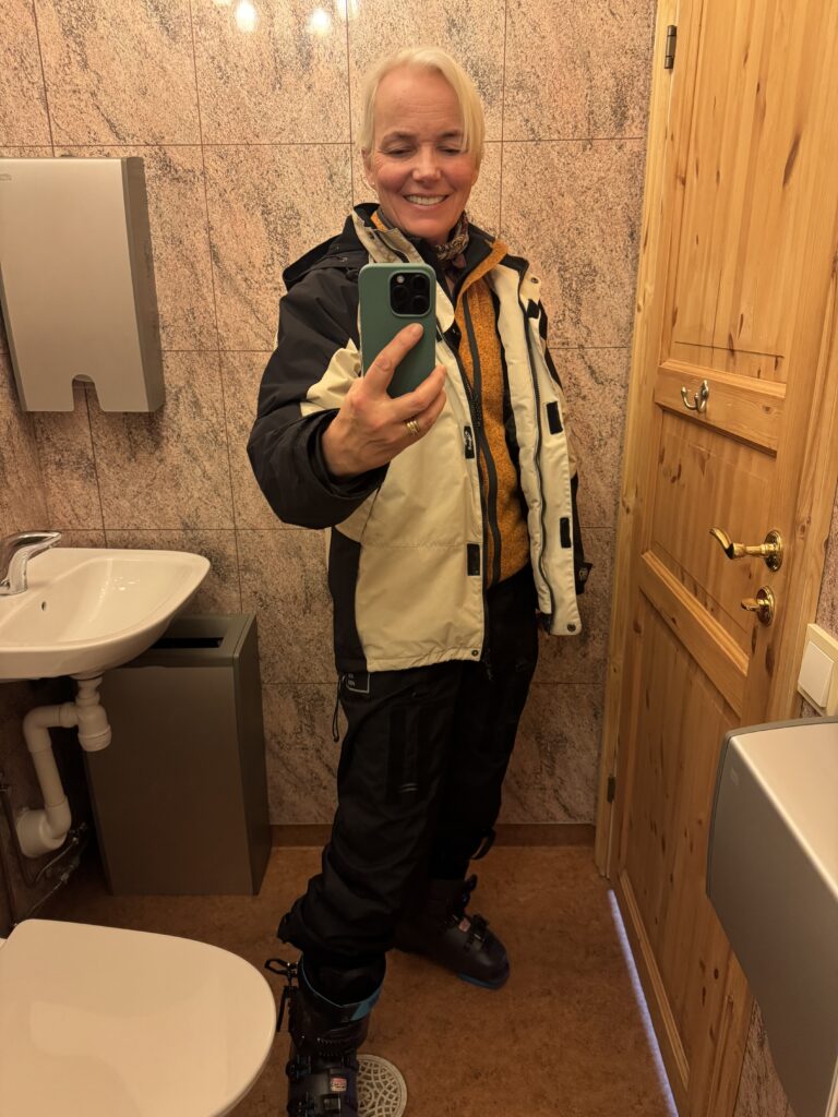 En person med kort blont hår tar en spegelselfie i ett badrum, iklädd en varm jacka, halsduk och skidbyxor med pjäxor, och ler mot kameran. Badrummet har ljusbruna väggar och trädörrar.