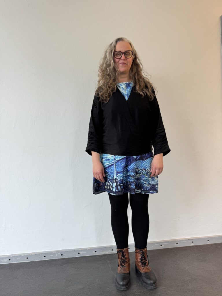 En kvinna med långt, lockigt grått hår och glasögon står inomhus mot en vit vägg. Hon är klädd i en svart topp, en blåmönstrad kjol, svarta leggings och bruna stövlar.