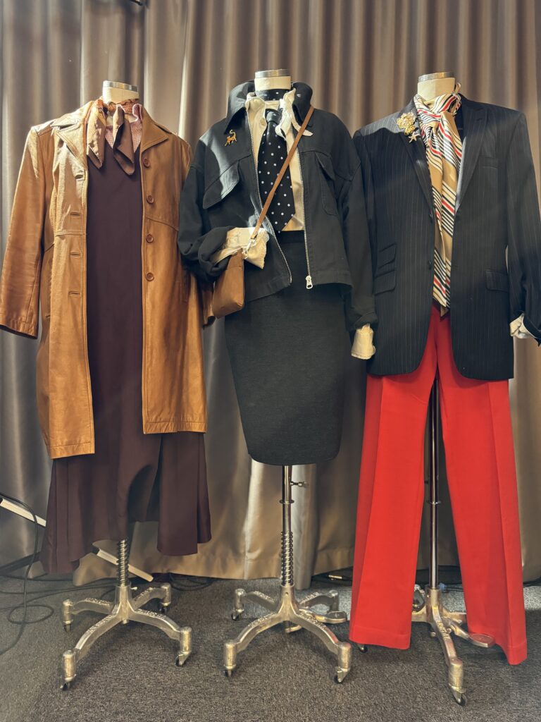 Tre skyltdockor klädda i vintagestil: en i brun klänning och solbrun kappa, en i svart kjol och kavaj med prickig slips och en i röda byxor, randig skjorta och mörk kavaj, alla står framför en gardin.