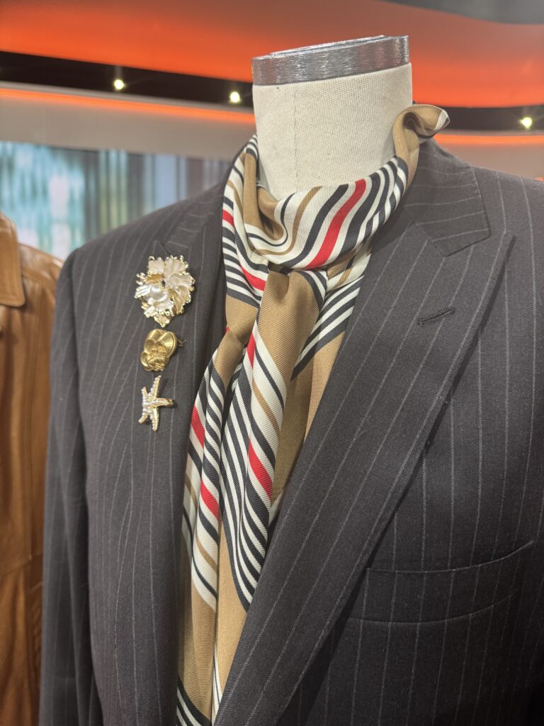 En skyltdocka klädd i en mörk kritstrecksrandig kostym med en brun, röd, vit och svartrandig halsduk instoppad. Tre dekorativa pins - en blomma, ett hjärta och en sjöstjärna - är fästa på kostymens kavajslag.