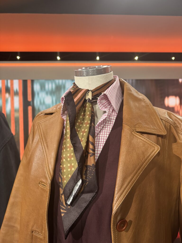 En skyltdocka visar en brun skinnrock över en vinröd tröja, en röd- och vitrutig skjorta och en mönstrad brun halsduk knuten runt halsen, mot en orange bakgrund.