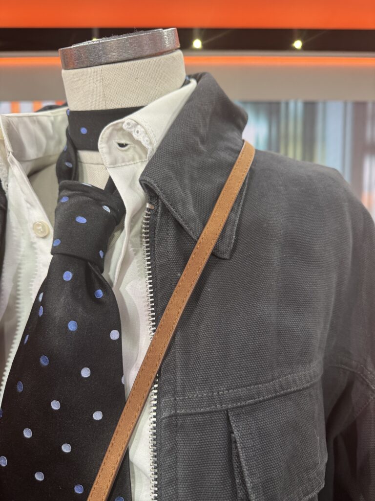 En skyltdocka klädd i vit skjorta med knappar, svart prickig slips och mörk kavaj med ett solbrunt band över axeln, visas inomhus med en suddig bakgrund.