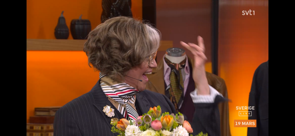 En leende person med kort, vågigt grått hår bär glasögon, kritstrecksrandig kostym och en randig scarf och håller i en färgglad bukett. Bakgrunden är orange med hyllor och en skyltdocka. TV grafik visar "SVT1" och "Sverige Live 19 mars.