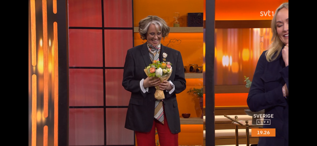 En leende person med grått hår, klädd i mörk kavaj, röda byxor och randig skjorta, håller i en blombukett på en starkt upplyst TV-apparat. En annan person syns delvis till höger. Texten på skärmen lyder "SVERIGE LIVE 19.26".
