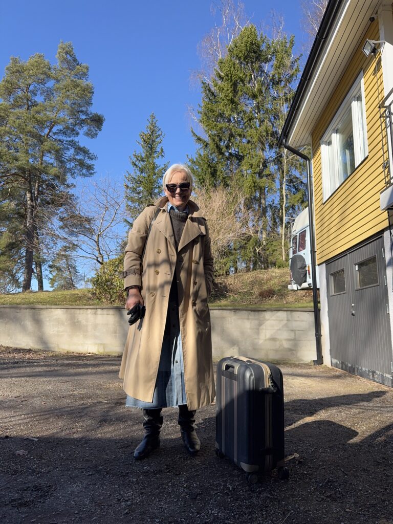 En äldre kvinna med solglasögon, beige trenchcoat och stövlar står utomhus med en resväska bredvid sig. Hon ler och i bakgrunden syns höga träd, ett gult hus och en blå himmel.