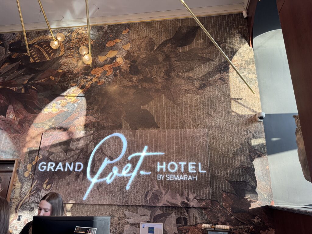 En neonskylt med texten "Grand Poet Hotel by Semarah" är monterad på en vägg med en blommönstrad väggmålning. Två personer sitter vid en receptionsdisk i det nedre vänstra hörnet, med solljus som strömmar in från höger.