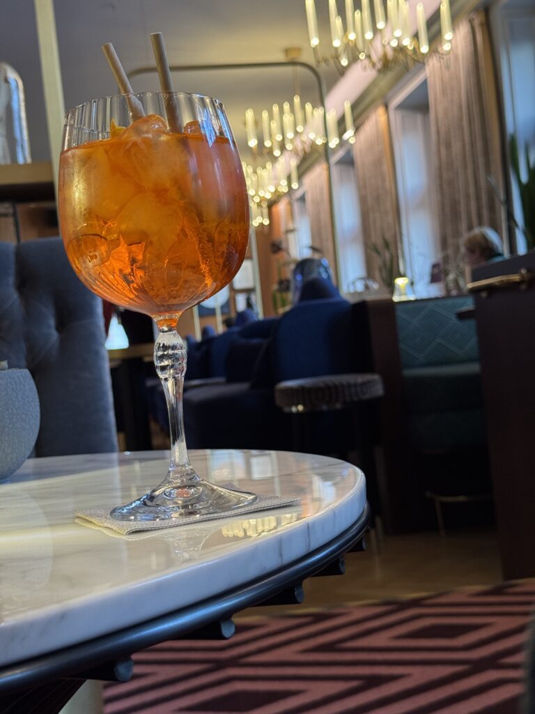 Ett stort vinglas fyllt med en orange cocktail och is står på ett marmorbord i en elegant lounge med modern belysning, plyschstolar och mönstrade mattor.