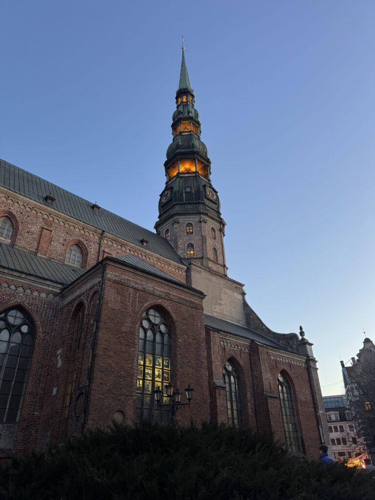 En hög, historisk tegelkyrka med grön spira och upplysta fönster i skymningen mot en klarblå himmel. Några byggnader och lövverk syns i förgrunden och bakgrunden.