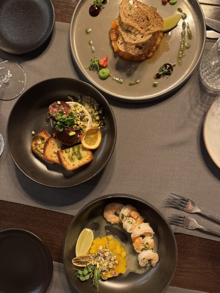 Tre tallrikar med eleganta gourmeträtter står uppdukade på ett bord, var och en konstfullt presenterad med garnityr, såsdroppar och rostat bröd eller bröd, tillsammans med bestick, tomma tallrikar och glas.
