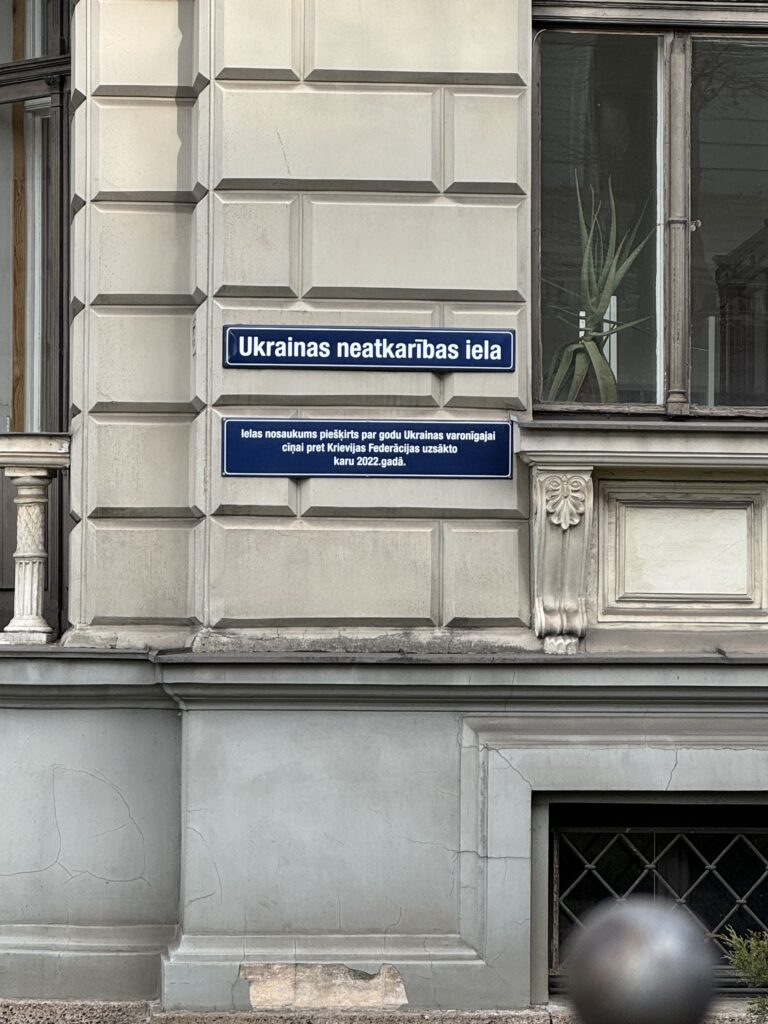 En husfasad med två blå gatuskyltar på lettiska; på den övre skylten står det "Ukrainas neatkarības iela" och på den nedre skylten förklaras att gatan är uppkallad för att hedra Ukrainas självständighet. En växt syns bakom ett fönster.