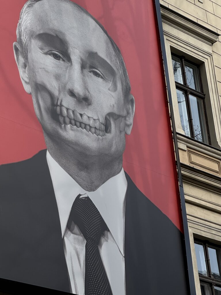 En skylt visar ett mansporträtt med en del av ansiktet digitalt förändrat för att avslöja en dödskalle under, mot en röd bakgrund, på sidan av en byggnad med fönster.