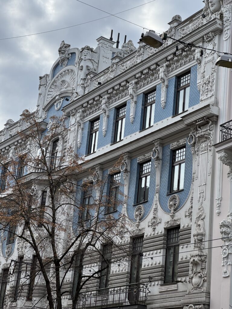 En utsmyckad byggnadsfasad med blå och vit dekorativ puts, inklusive skulpterade ansikten, intrikata mönster och stora fönster; ett lövlöst träd står framför under en molnig himmel.