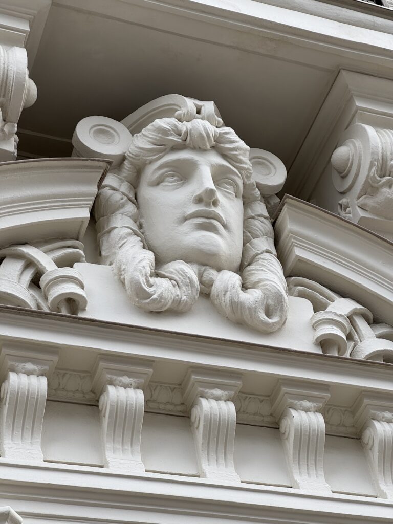 Utsmyckad vit arkitektonisk detalj med ett skulpterat ansikte med långt, vågigt hår omgivet av dekorativa element på en klassisk byggnadsfasad.