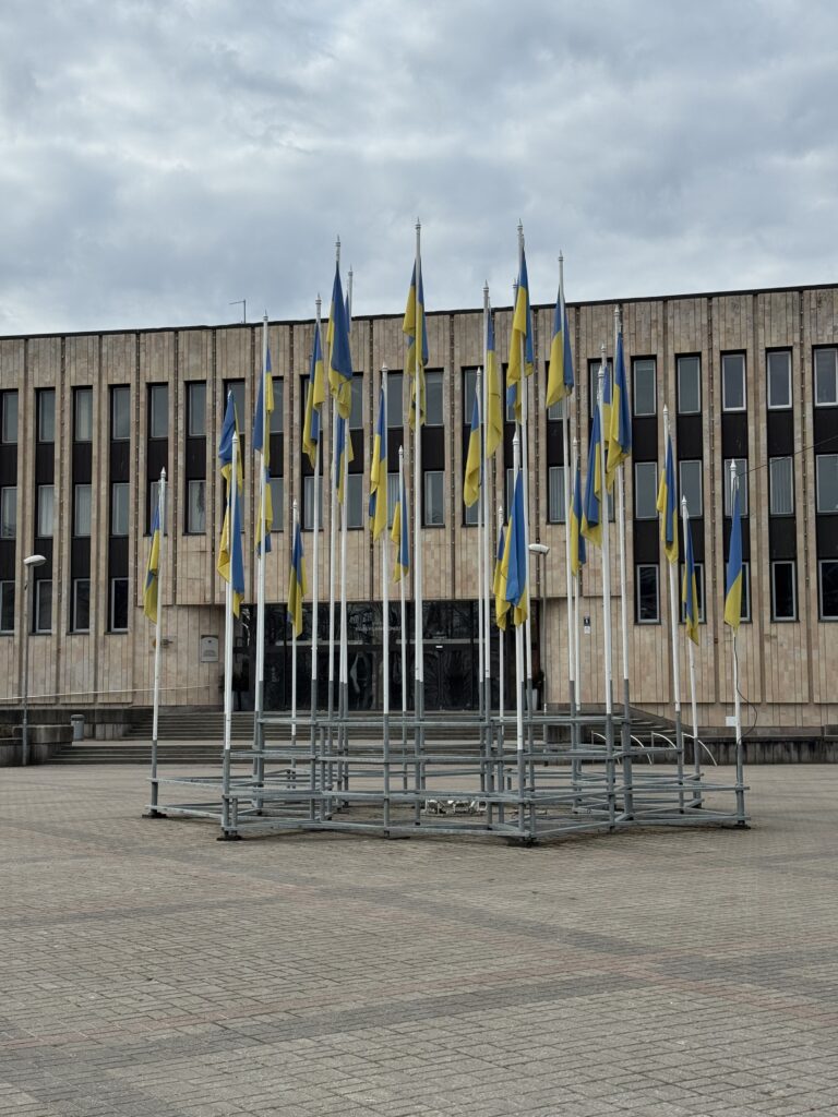 Flera ukrainska flaggor är grupperade på flaggstänger framför en stor, rektangulär byggnad med många fönster och en stenfasad under en molnig himmel.
