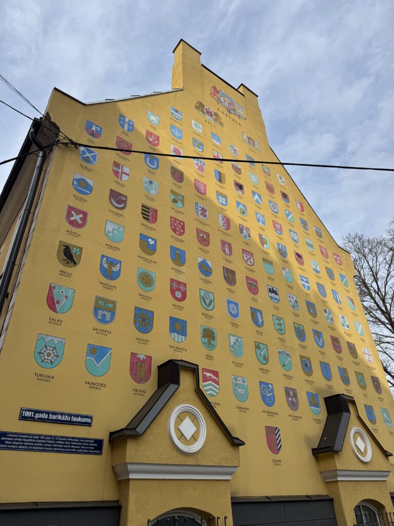 En hög gul byggnad med dussintals färgglada vapensköldar i rader över fasaden, med en blå himmel ovanför och några nakna trädgrenar på höger sida.