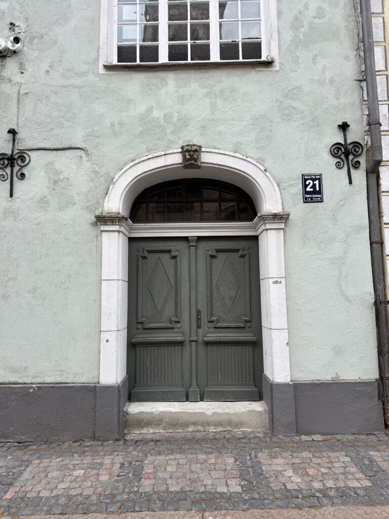 En grå entré med dubbeldörr med dekorativa lister och ett litet välvt fönster ovanför, i en ljusgrön byggnad. En skylt med gatunumret "21" syns på väggen. Trottoaren är belagd med kullerstenar.