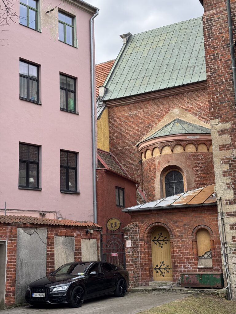 En svart bil står parkerad framför en rosa och en röd tegelbyggnad med välvda fönster och dörrar. Byggnaderna har olika arkitektoniska stilar och väderbitna texturer. Himlen är mulen.