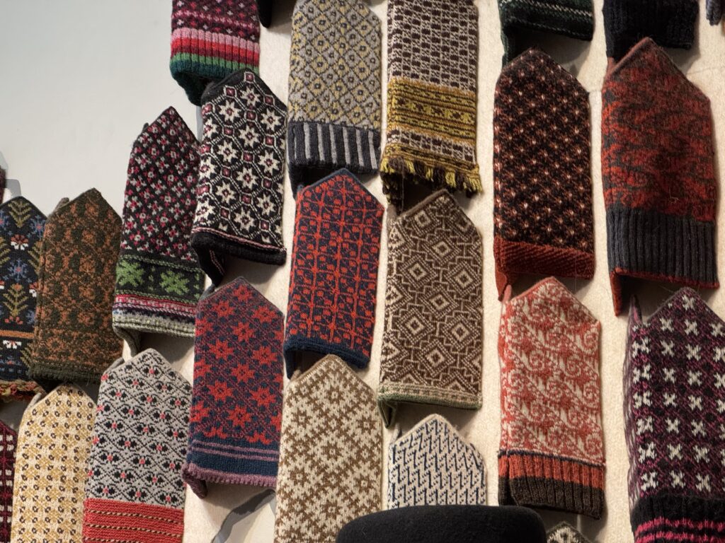 En uppvisning av färgglada, intrikat mönstrade stickade vantar i rader på en vägg, med olika traditionella mönster och livfulla kombinationer av rött, svart, vitt, brunt och andra färger.