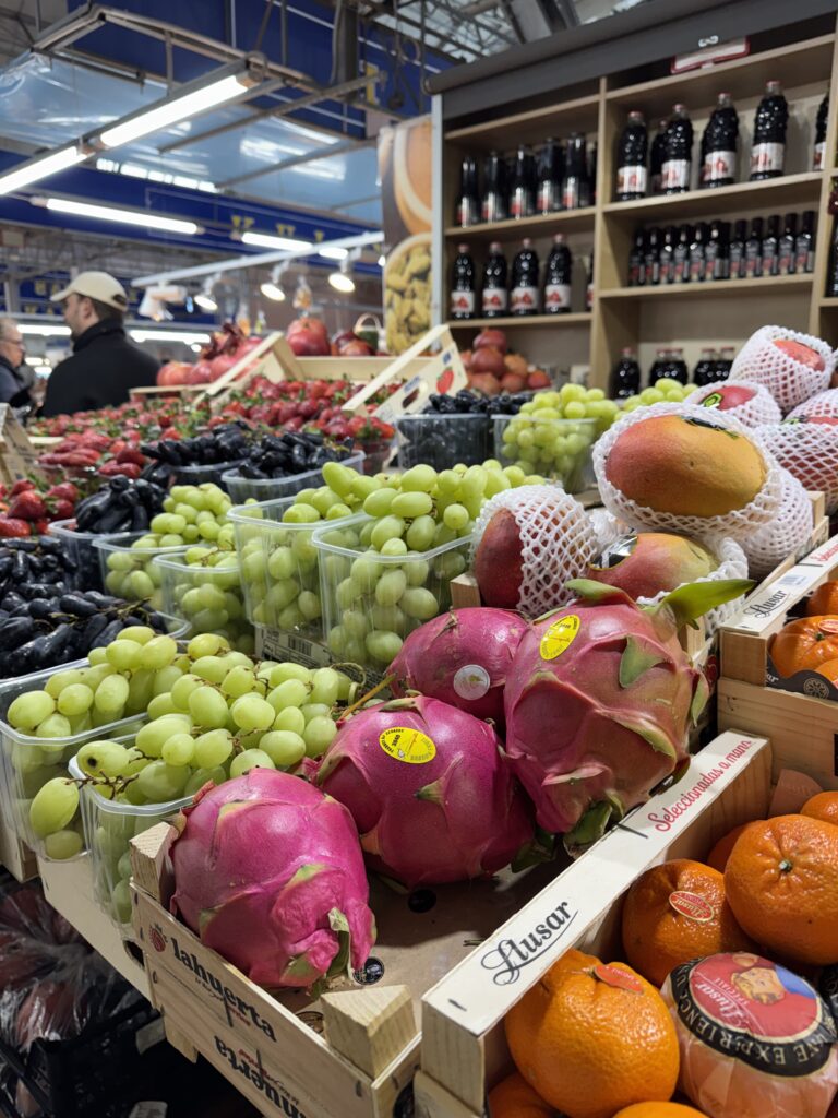 Färgglad fruktdisplay i en livsmedelsbutik med drakfrukt, gröna druvor, mango, apelsiner och andra frukter. Hyllor med flaskdrycker syns i bakgrunden, tillsammans med shoppare.