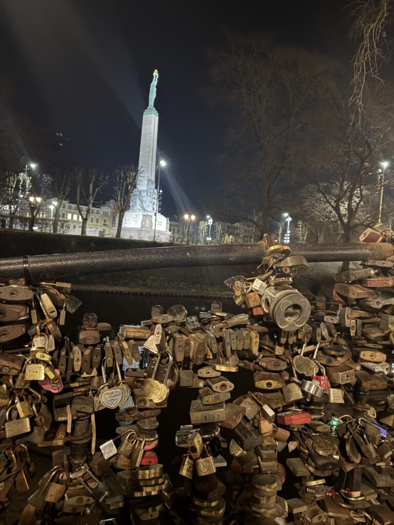 En nattlig vy av ett stängsel täckt av hänglås, med det upplysta Frihetsmonumentet i bakgrunden, omgivet av träd och stadsljus.