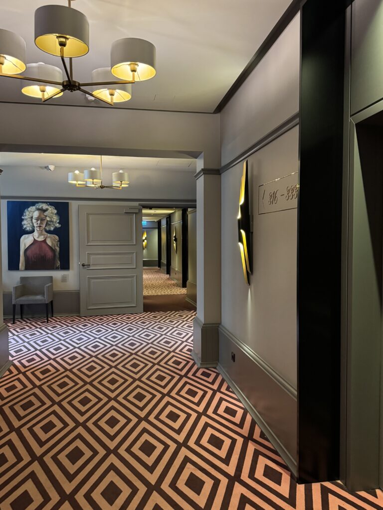 En hotellkorridor med geometriskt mönstrad matta, moderna armaturer och rumsskyltar. Ett stort porträtt av en kvinna hänger på väggen nära en stol. Flera dörrar och en spegel syns längs korridoren.