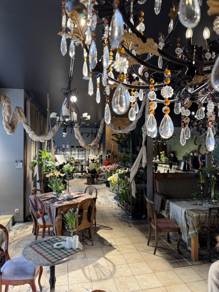 Ett mysigt café med vintagemöbler, hängande kristallkronor, krukväxter på borden, draperad tygdekoration och en barista som arbetar bakom disken i bakgrunden.