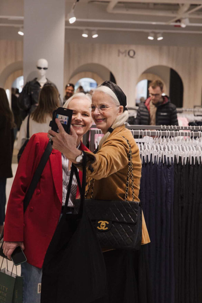 Två äldre kvinnor ler och tar en selfie tillsammans i en klädaffär, stående framför ställningar med kläder. Andra shoppare tittar i bakgrunden. Den ena kvinnan bär en röd kavaj, den andra en brun kappa med svart pannband och Chanelväska.