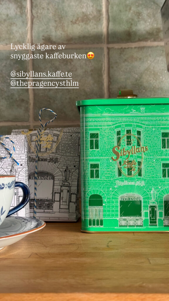 En grön Sibyllans kaffeburk med en illustrerad byggnad ligger på en bänkskiva av trä bredvid en blåvit tekopp och en gråmönstrad låda. Text på svenska är inlagd överst.
