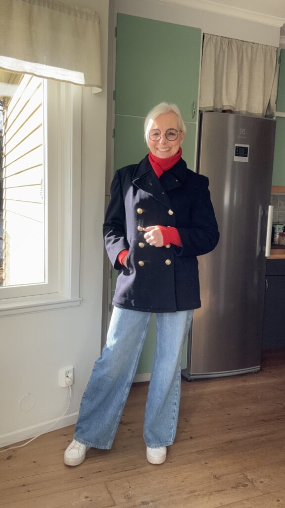 En äldre kvinna med vitt hår och glasögon står och ler inomhus. Hon bär en svart dubbelknäppt kappa, röd polotröja, vida blå jeans och vita sneakers. Hon står på ett trägolv nära ett fönster och ett grönt kök.