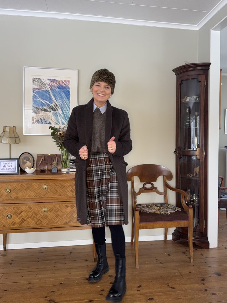 En person ler inomhus, klädd i en brun kappa, rutig kjol, mörka strumpbyxor, svarta stövlar och en hatt med leopardmönster. De står i ett mysigt, traditionellt inrett rum med trämöbler och inramad konst på väggen.