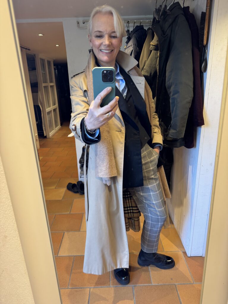 En person med kort vitt hår tar en spegelselfie och ler medan han bär en beige trenchcoat, blå skjorta, svart kavaj, rutiga byxor och svarta skor. Kapporna hänger på krokar bakom dem i en hall med klinkergolv.