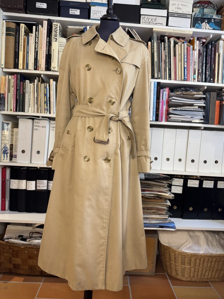 En lång beige trenchcoat med bälte visas på en svart skyltdocka framför hyllor fyllda med böcker, mappar och staplade papper i en studie- eller kontorsmiljö.