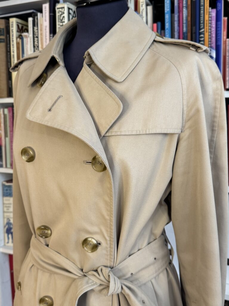 En beige dubbelknäppt trenchcoat med bälte visas på en skyltdocka framför en bokhylla fylld med olika böcker.