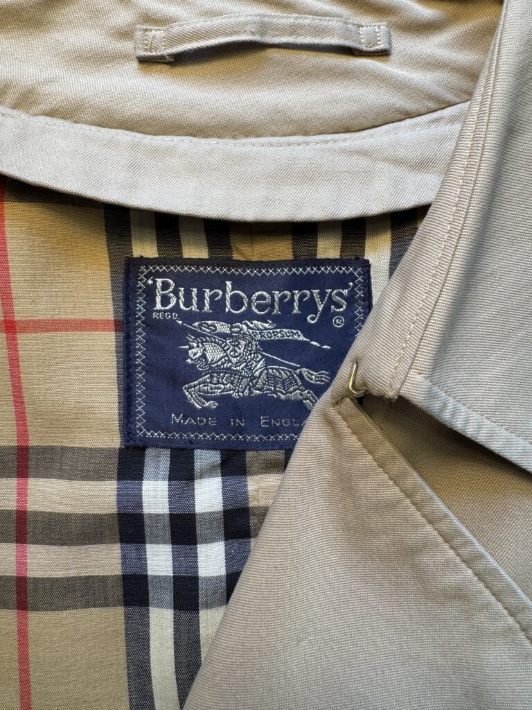 Närbild av en beige Burberrys-skjorta med uppvikt krage, med en marinblå etikett med texten "Burberrys Made in England" och Burberrys karakteristiska rutmönster i beige, svart, vitt och rött.