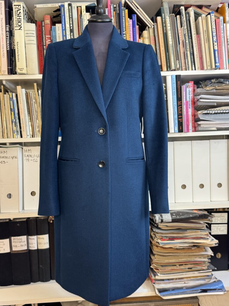 En marinblå skräddarsydd kappa med två knappar visas på en skyltdocka framför hyllor fyllda med böcker, tidskrifter och mappar.