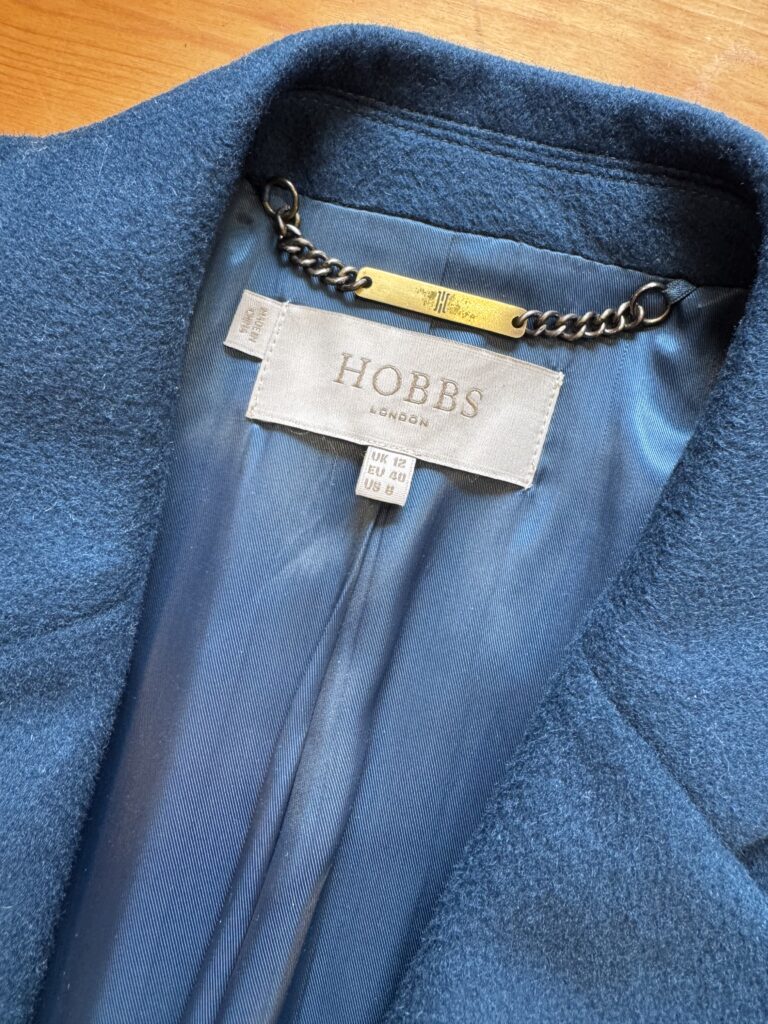 Närbild på insidan av kragen på en blå Hobbs London-kappa, med märkesetikett, en namnskylt i guld fäst vid en svart kedja och en storleksetikett som anger UK 12, EU 40 och US 8.