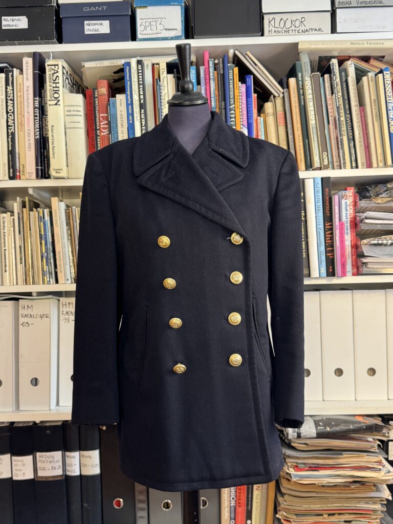 En svart dubbelknäppt kappa med guldknappar visas på en klädform framför hyllor fyllda med böcker, pärmar och tidskrifter.
