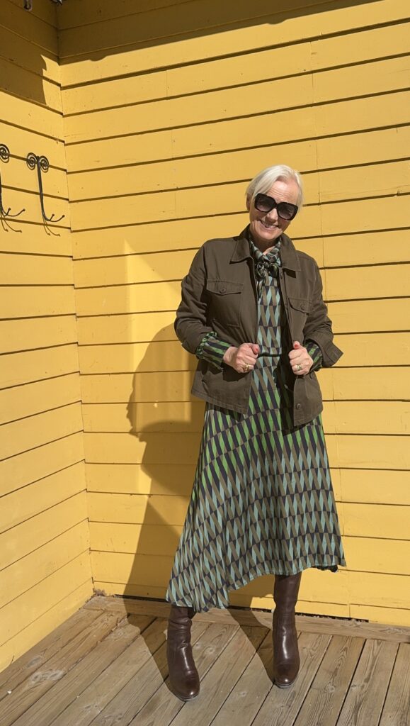 En kvinna med kort vitt hår och solglasögon står mot en ljusgul trävägg, iklädd en mönstrad grön och blå klänning, en mörkgrön jacka och bruna stövlar. Solljuset kastar en skugga bakom henne på däcket.