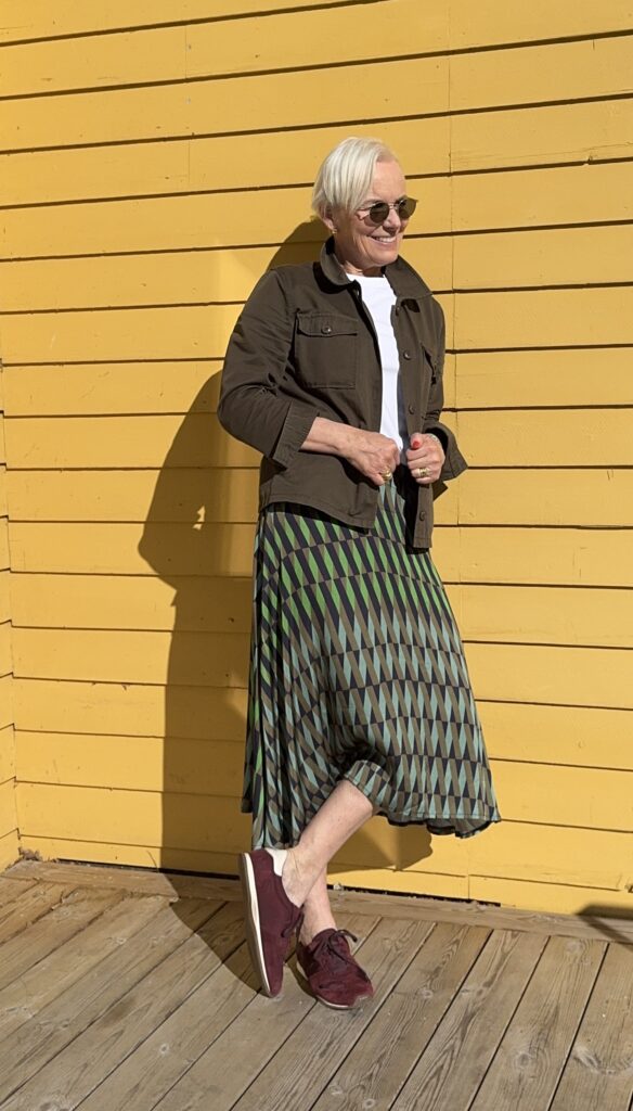 En person med kort vitt hår, solglasögon och mörk jacka står leende mot en ljusgul trävägg, iklädd en geometriskt mönstrad kjol och rödbruna sneakers.
