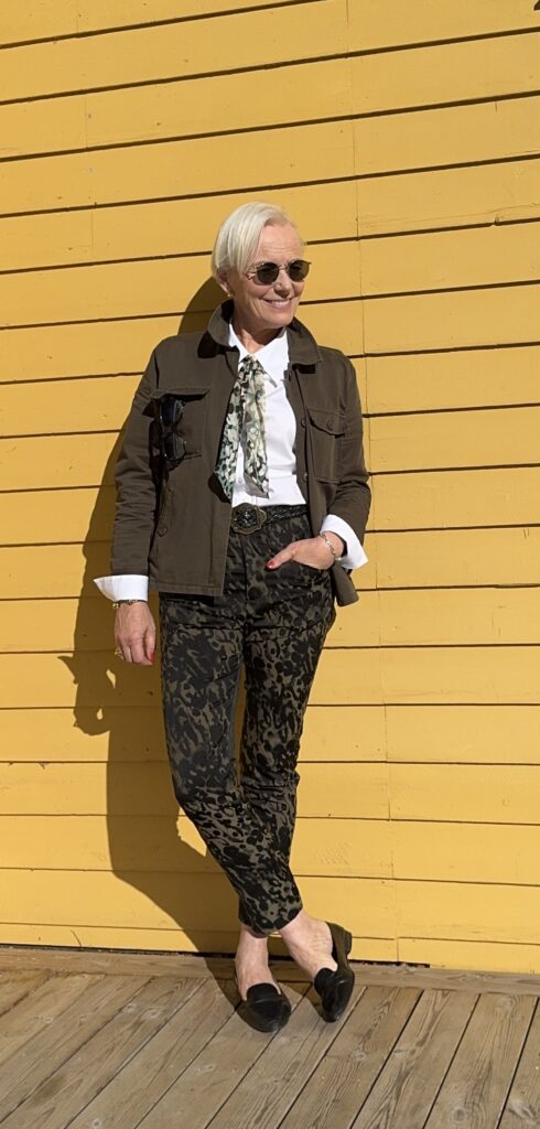 En elegant äldre kvinna med kort blont hår och solglasögon lutar sig mot en ljusgul trävägg. Hon bär en mörk jacka, vit skjorta, mönstrad scarf, leopardmönstrade byxor och svarta skor och ler med armarna i kors.